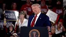 Politiker Donald Trump i kostym håller tal vid podium med presidentens sigill, publik i bakgrunden.