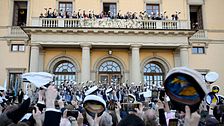 Studenter firar utanför skolbyggnad, viftar med mössor mot balkong.
