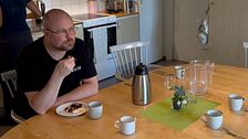 Anton Bylund har svarta kläder med Skellefteå kommuns logga på bröstet. Han sitter vid ett bord uppdukat med kaffekoppar med en bit kladdkaka i handen.