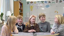 Fem barn sitter vid ett bord i ett klassrum och skrattar. I bakgrunden syns en whiteboardtavla och en bokhylla.