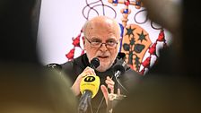 Presskonferens med mikrofoner och vapensköld i bakgrunden.