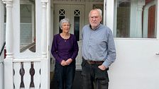 En kvinna och en man står utanför en vit verandadörr, kvinnan i lila tröja, mannen i blå skjorta.