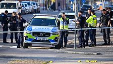 Poliser vid avspärrning med polisbil på stadsgata, flera personer i bakgrunden.