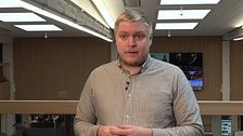 En person med randig skjorta står inomhus framför en träpanel och stora lampor.
