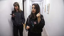 Nooshi Dadgo stands in a corridor, talking and holding up his hand while a woman stands behind him with a cell phone.