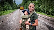 En kvinna står med en bebis i bärsele på magen framför en brandbil