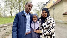 Tre personer står tillsammans utomhus, med stenvägg och byggnad i bakgrunden.