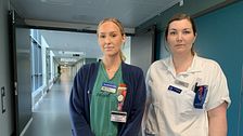 Två sjuksköterskor står i en korridor. Kvinnan till höger har vita kläder och kvinnan till vänster har grön tröja och blå kofta över.