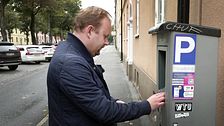 En person står vid en parkeringsautomat och använder den, med ett parkeringstecken synligt på automaten. I bakgrunden syns en rad med parkerade bilar och en gata kantad av träd.