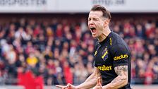 Fotbollsspelare i svart tröja firar framför publik på läktaren.