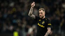Fotbollsspelare i AIK-tröja höjer handen under match, suddiga åskådare i bakgrunden.