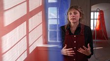 Person gestikulerar i rödbrun skuggljus interiör med färgglad vägg och fönster.