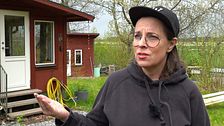 Person med keps utanför ett rött hus med trädgårdsslang och grönska i bakgrunden.