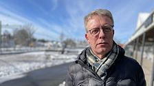 Per Högberg (KD) i vinterjacka står vid snötäckt park, med en solig himmel och promenadstråk i bakgrunden.