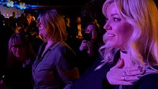 en blond kvinna som ler och håller sig över bröstet. i ett publikhav.