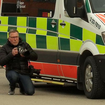 Fotografen Öyvind Lund står på knä bredvid en sjuktransportbil och fotograferar.