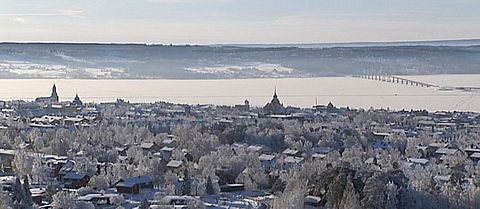 Vy över Östersund täckt av snö med dimma över sjö och Vallsundsbron i bakgrunden.