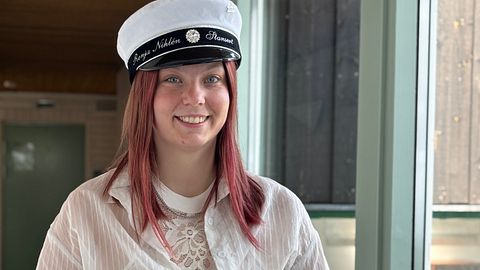 Student med vit mössa och ljust hår står inomhus vid ett fönster.