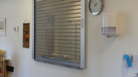 På bilden syns avdelningsköket bakom en gallerliknande jalusi. På väggen intill finns en bonad, en klocka och en flaska med desinfektionsmedel.