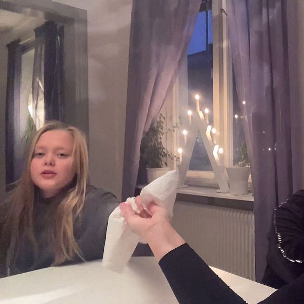 Två barn och en vuxen person sitter vid ett bord. Den vuxna personen räcker en näsduk till det ena barnet.  I fönstret bakom bordet står en adventsljusstake.