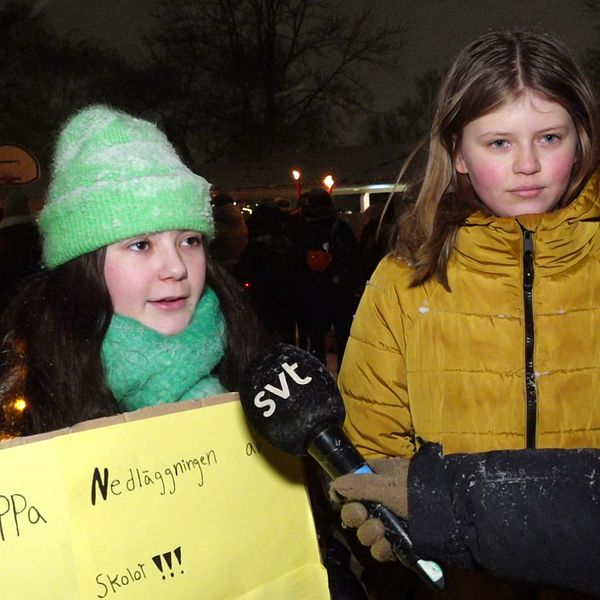 Till vänster står två flickor, en med mörkt hår, grön halsduk och mössa och en gul skylt som det står stoppa nedläggningen på. Den andra flickan har en gul jacka på sig. Till höger syns en grupp människor samlade