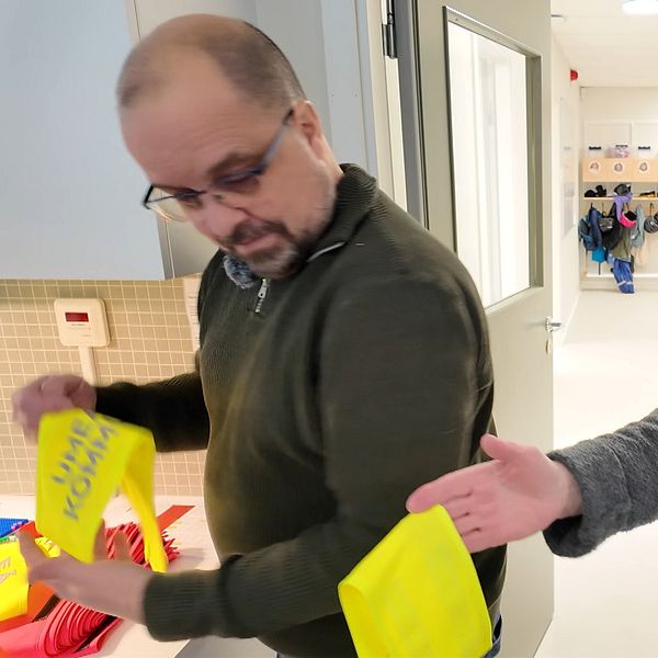 Två personer står i en ljus korridor med dörröppningar i bakgrunden. Den manliga personen till vänster håller en gul armbindel, medan den kvinnliga personen till höger sträcker fram en annan gul armbindel. Korridoren är inredd med vita väggar och det syns klädhängare med flerfärgade kläder i bakgrunden.