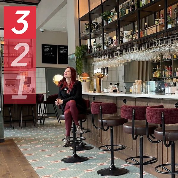 En stilren bar med en lång bänkdisk, inredd med trä och metall. Hyllor med alkoholflaskor syns bakom disken, där glas hänger i rader. Emma Engdahl, kärleksexpert, sitter på en barstol klädd i svarta kläder, vänd mot kameran. Golvet är dekorerat med ett mönstrat kakel och det finns växter i lokalen som bidrar till den moderna atmosfären.