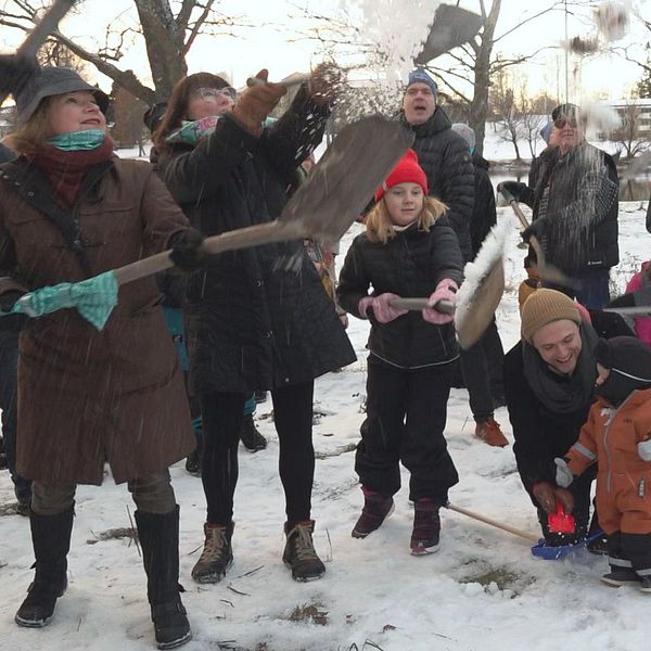 En grupp människor, inklusive barn, deltar entusiastiskt i en snöbollskastning eller lek i ett snötäckt parkområde. Några i gruppen använder spadar för att hantera snön. Stämningen verkar glad och livlig med en bakgrund av träd och en byggnad i fjärran.