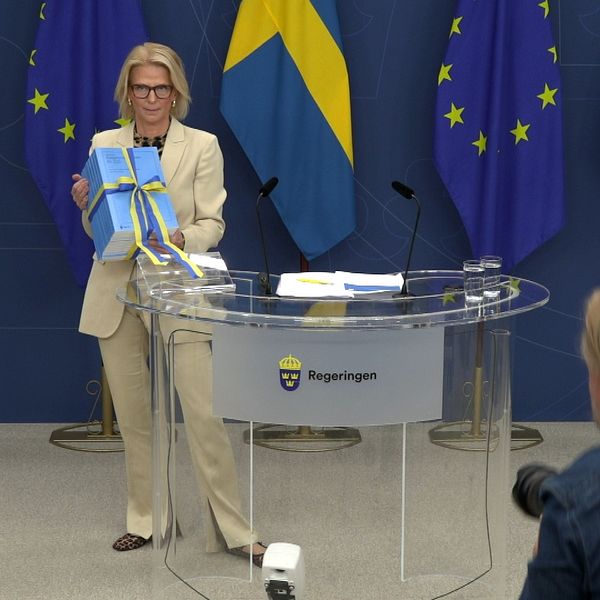 En delad bild som visar två scener. Till vänster står en person vid en presskonferens med en present insvept i blått papper och ett band. I bakgrunden syns svenska och europeiska flaggor. Till höger är en nattbild av en tågstation med upplysta byggnader och spår, vilket skapar en lugn atmosfär.