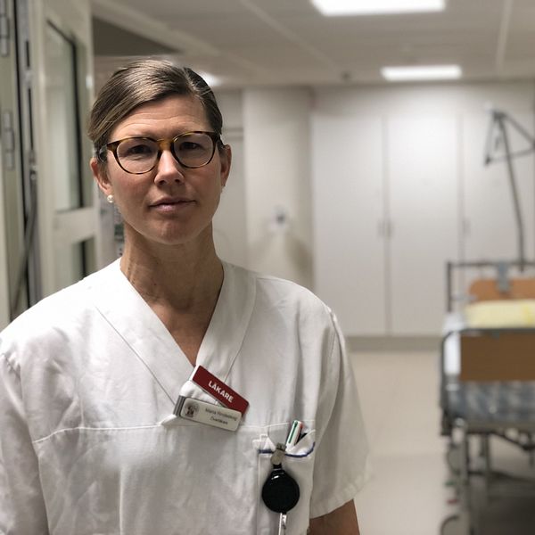 En läkare i vit uniform står i en sjukhuskorridor. Bakgrunden visar en tom sjukhussäng med sängkläder, vilket indikerar en läkarmiljö.