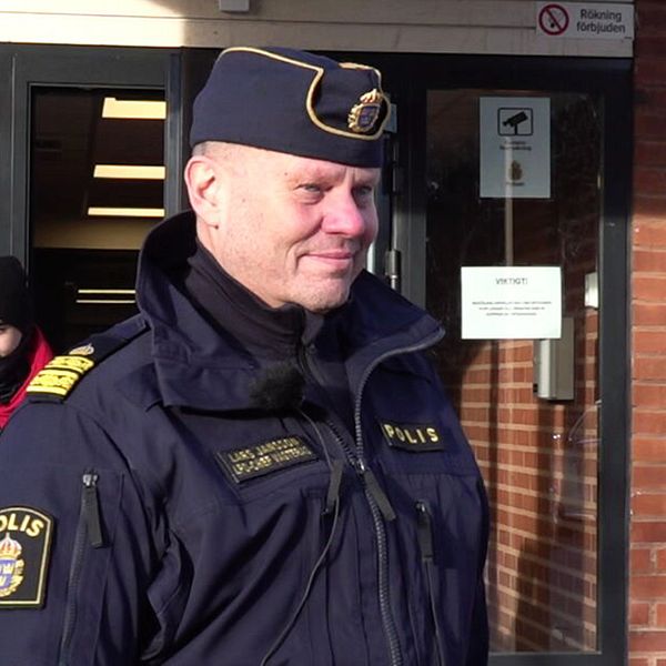 En polis står utanför en byggnad och samtalar med en annan person. Bakgrunden visar en ingång med skyltar och fönster. Den polis uniformen syns tydligt, vilket indikerar en officiell situation.
