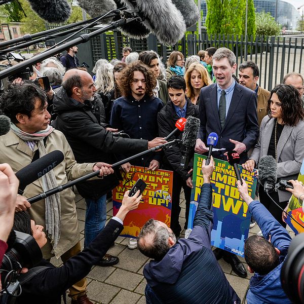 Unga människor omringade av journalisters mikrofoner, en äldre dam i grått kort hår och pärlhalsband