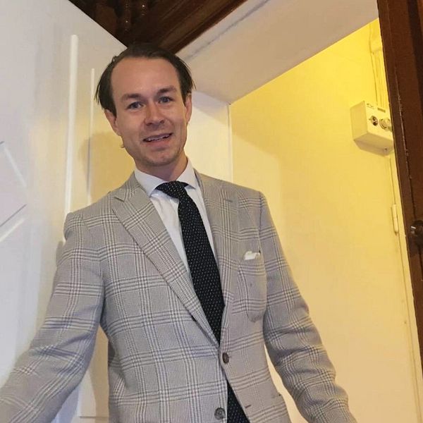 En delad bild som visar två sektioner. Till vänster står en man i kostym vid en dörröppning i en ljus korridor. Till höger visas en stilren lägenhet med trägolv, ett litet matbord med stol och en vit byrå, samt fönster som släpper in naturligt ljus.