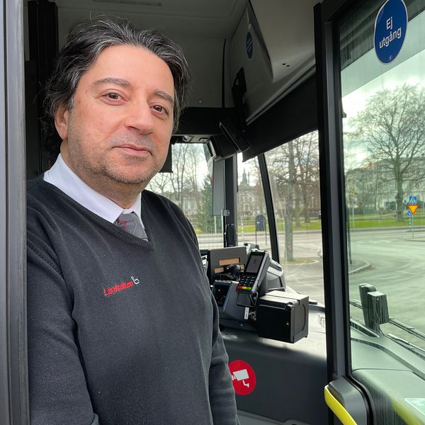 En delad bild: Till vänster står en person i en buss, klädd i en arbetsuniform med företagslogotyp på tröjan. Till höger vajar en svensk flagga på halv stång framför en vit byggnad, kanske en kommun- eller statlig fastighet. Det är en mulen dag med träd utan löv i bakgrunden.