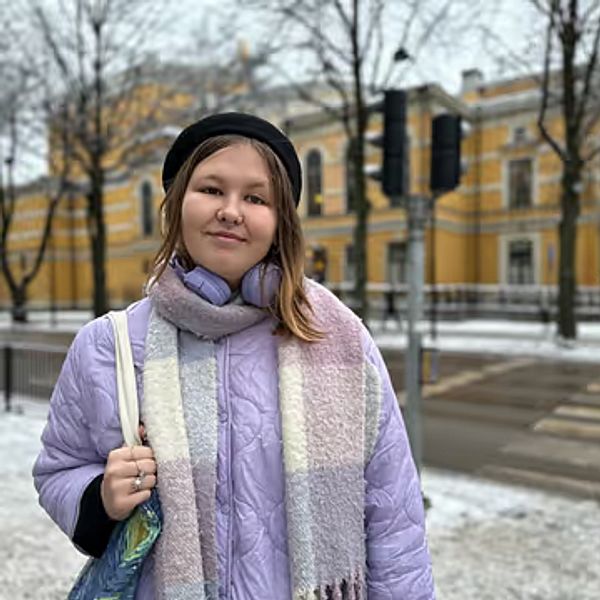Två personer står utomhus på en snötäckt gata. Den vänstra personen bär en lila jacka och en scarf, medan den högra personen har på sig en camouflage-mönstrad jacka med en huva. I bakgrunden syns en byggnad med gul fasad och trädkronor utan löv.
