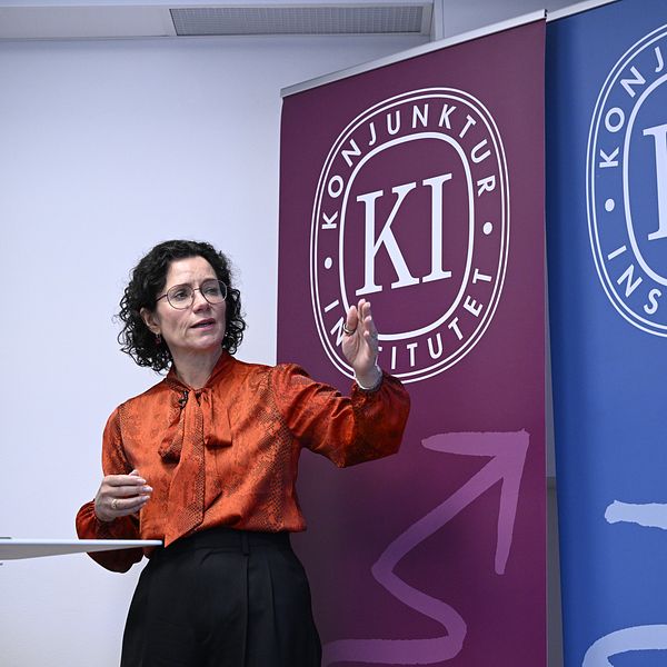 En person står och presenterar framför två banner med logotyper för Konjunkturinstitutet, en i röd och en i blå färg.