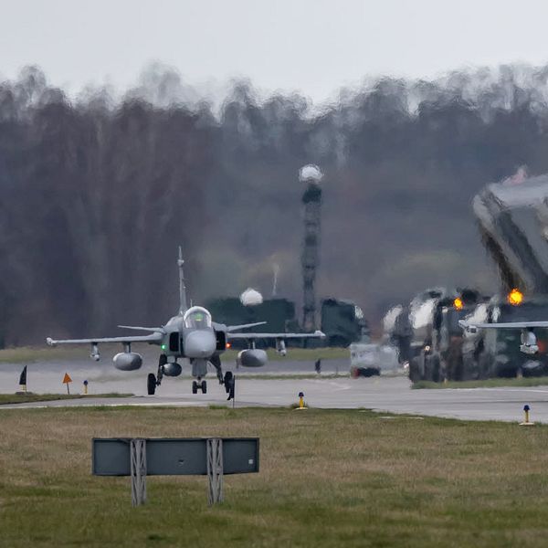 Två stridsflygplan taxiing på en flygbas med hangarer i bakgrunden.