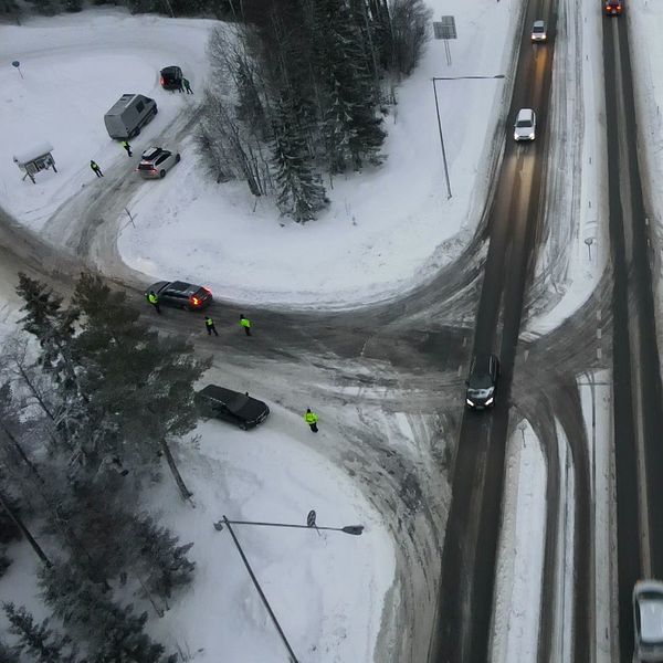Vy av en trafikerad korsning under vinterförhållanden, där snö täcker vägen och omgivande landskap. Flera fordon rör sig genom korsningen, och två personer i reflexkläder står vid sidan av vägen. Området omges av träd och en sjö syns i bakgrunden.
