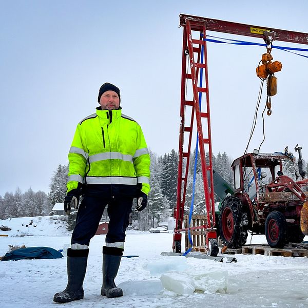 Man i gul varseljacka står på Näktens is bredvid en traktor som bärgats från sjöbottnen.