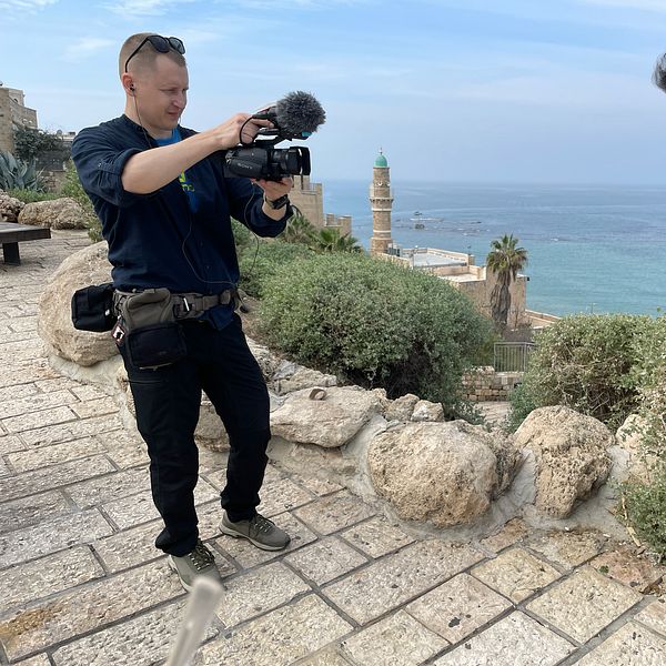 Samir Abu Eid står på en stenlagd promenad med utsikt över havet, medan Johannes Tolf filmar med en kamera. I bakgrunden syns en historisk byggnad och en utsiktstorn, med palmer och havet i horisonten.