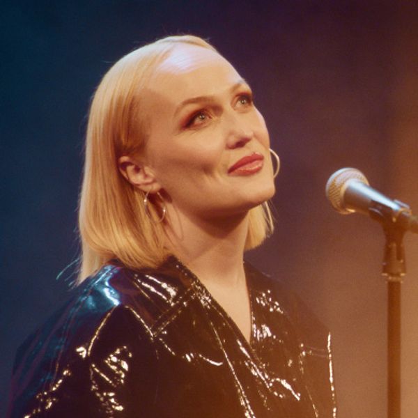 En kvinna iklädd svart lackjacka med kort, blond page står framför en mikrofon
