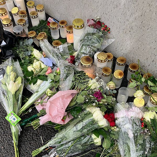 Till vänster syns en minnesplats med blommor och tända ljus, som hedrar en avliden person. Till höger står en reporter i en korridor med ett mikrofoninspelning från en nyhetskanal.