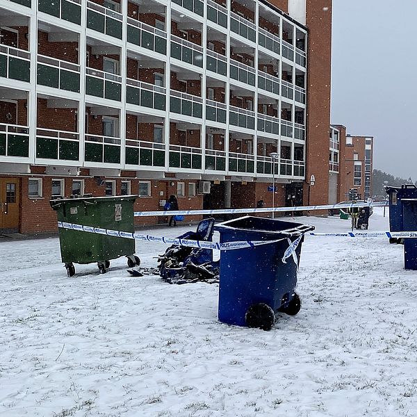 Sju soptunnor i olika färger, inklusive gröna och blå, står på en snötäckt gräsmatta framför en flerfamiljsbyggnad i stadsdelen Lugnvik i Östersund. Området är avspärrat med blå-vita polisband, och snö faller lätt. Bakom byggnaden syns träd i bakgrunden.