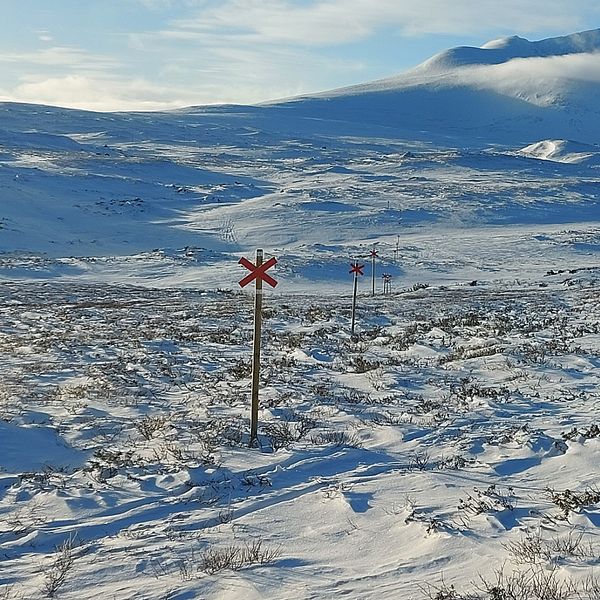 Vy mot Helagsmassivet. Tunt snötäcke. På bilden synsett rött ledkryss som markerar en vinterled i förgrunden och bergen i bakgrunden under en klarblå himmel.