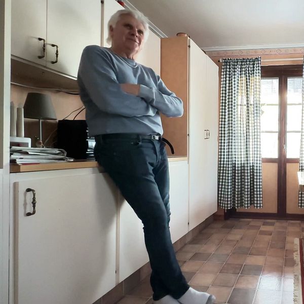 Köksmiljö med ett matbord och stolar i trä, samt ett fönster med rutiga gardiner. I bilden syns en person som lutar sig mot en köksskåp. På väggen hänger några tavlor. Golvet är beklätt med plattor och en matta ligger under matbordet.