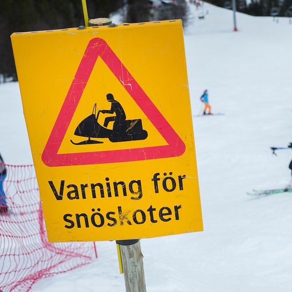Skidbacke med varningsskylt för snöskoter, skidåkare i bakgrunden.