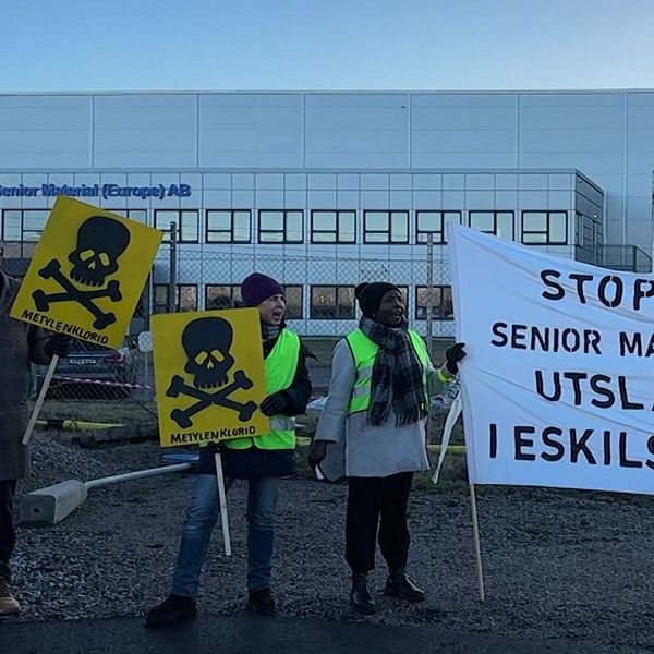 Grupp människor i gula reflexvästar håller skyltar och en banderoll framför en industribyggnad som tillhör Senior Material (Europe) AB. Banderollen har texten ”STOPPA SENIOR MATERIALS UTSLÄPP I ESKILSTUNA” och uppmanar till åtgärder mot miljöutsläpp. Några skyltar visar bilder av dödskallar för att varna för fara.