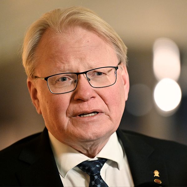 En delad bild som visar två personer i formella kläder. Till vänster syns en man med ljus färg på håret och en mörk kostym, medan han, tillsammans med andra, deltar i en offentlig verksamhet. Till höger är en annan man klädd i en blå kostym med en ljus skjorta och ett mönstrat näsduk. Bakgrunden är suddig och rör sig om en scen med europeiska flaggor.