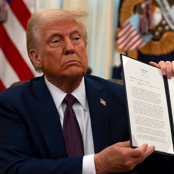 Donald Trump iklädd kostym håller upp ett dokument med en stor underskrift i ett formellt rum med gulddraperier och den amerikanska flaggan i bakgrunden.