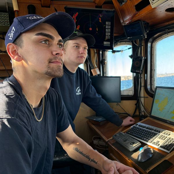 Två personer navigerar i en skeppshytt med havsutsikt och radarutrustning.
